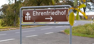 Besucherinnen und Besuchern wird an der Straße durch ein Hinweisschuld der Weg zur Ehrengräberstätte gewiesen.