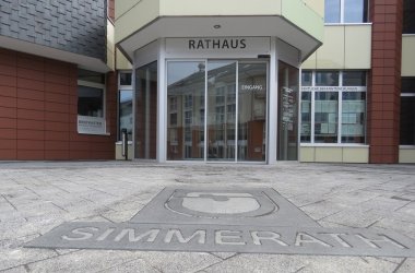 Der Eingang des Rathauses aus der Froschperspektive, im Vordergrund sind ein Pflasterstein mit dem Schriftzug "Simmerath" und dem Simmerath Wappen zu sehen